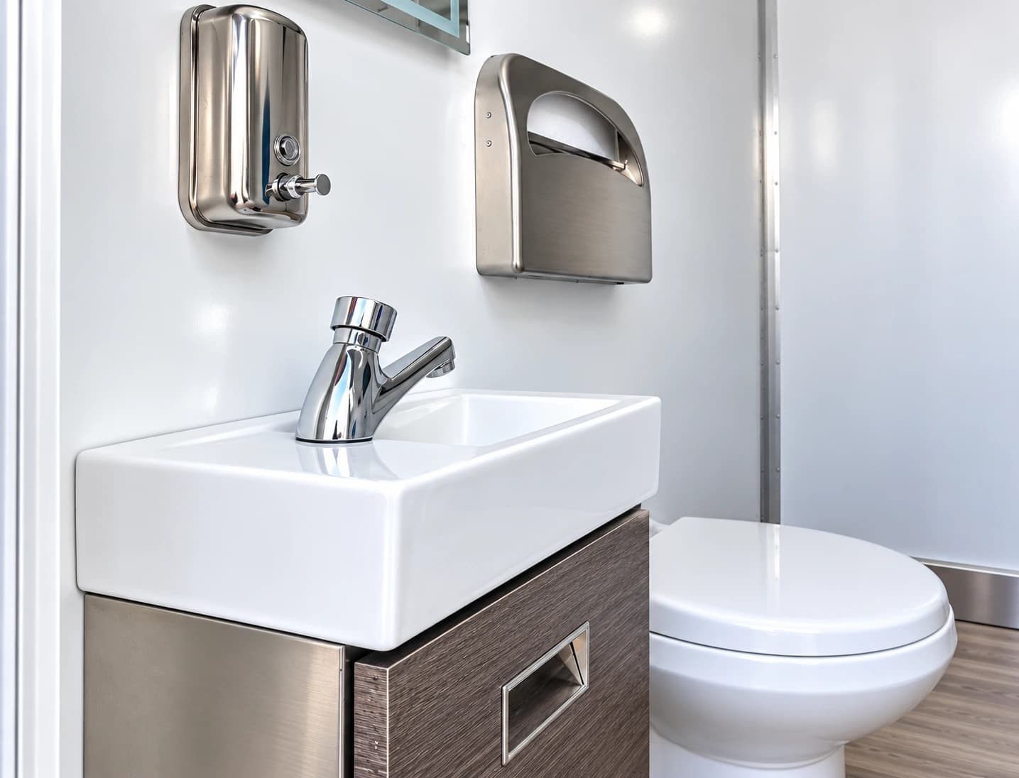 Small handwashing sink and wall-mounted dispensers beside a toilet inside a restroom trailer